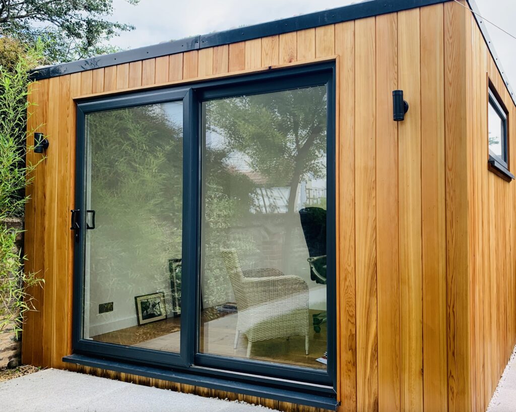Contemporary wooden garden room with large sliding glass doors and modern black frame, surrounded by greenery